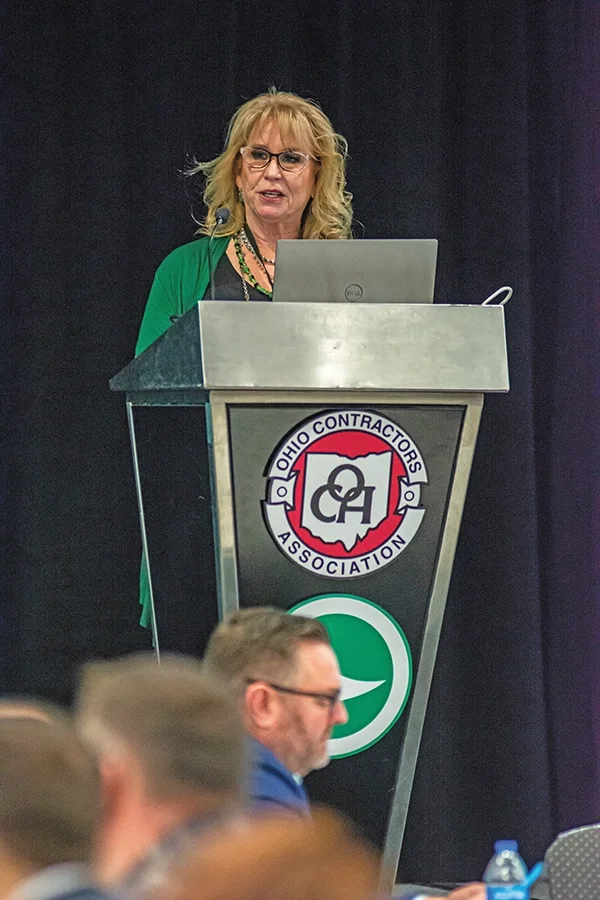 Female speaker at a metal podium with laptop, presenting at the Ohio Contractors Association event; audience seated.