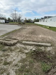 A vacant dirt lot with gravel and curb blocks beside a residential street; a small excavator sits in the distance.