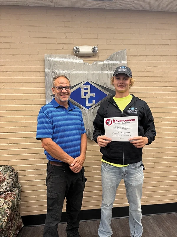 Two men indoors by a brick wall; one holds an Advancement Foundation certificate, with a blue diamond emblem behind.